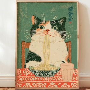 Op de afbeelding: Een rechthoekige wandkunstprint met een zwart-witte kat die aan een tafel zit en noedels eet uit een kom met patronen. De kat staat tegen een groene achtergrond met Japanse tekst en de tafel is bedekt met een rood kleed met witte bloemmotieven.