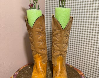 May include: A pair of tan leather cowboy boots with decorative stitching on the shafts and pointed toes. Each boot contains a bright green foam cylinder holding a bouquet of dark red chrysanthemum flowers.
