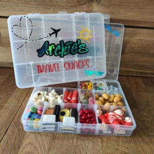 May include: A clear plastic snack box, divided into compartments, filled with sweets and snacks. The lid features a heart, airplane, sun, and the text "Archie's Travel Snacks."