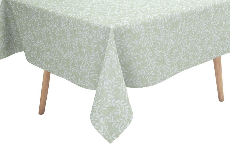 May include: A rectangular tablecloth in a soft sage green color featuring a repeating white botanical leaf pattern. The fabric drapes over the edges of a table with light-colored wooden legs, showcasing a consistent print across the entire surface.