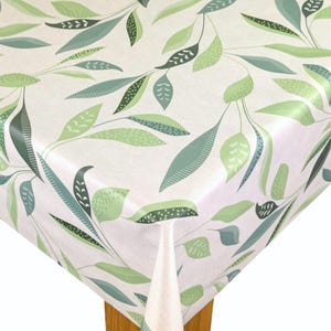 May include: A white tablecloth featuring a repeating pattern of green and teal leaves and stems. The leaves vary in size and shape, with some featuring dotted and striped details. The tablecloth is draped over a wooden table.