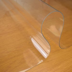May include: Clear plastic table protector rolled up on a wooden surface. The protector is designed to protect tabletops from scratches and spills.