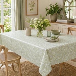 May include: A rectangular tablecloth in a soft sage green hue featuring a delicate, repeating white botanical leaf pattern. The fabric drapes over a dining table, providing a neutral base for a clear glass vase filled with white flowers.