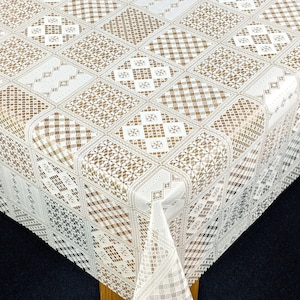May include: A white and beige tablecloth with a geometric design. The tablecloth has a repeating pattern of squares and diamonds, each filled with different patterns such as crosshatch, floral, and geometric shapes. The tablecloth is made of a shiny material.