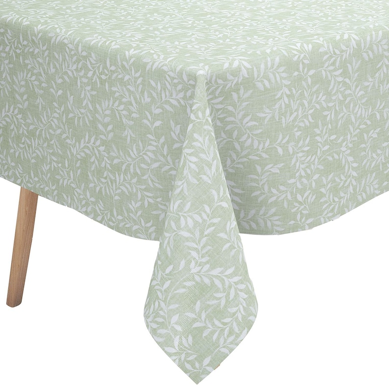 May include: A sage green tablecloth featuring a repeating white botanical leaf pattern. The fabric has a visible woven texture and drapes over the corner of a table with a light-colored wooden leg visible underneath the draped corner of the cloth.