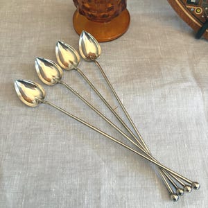 May include: Four silver cocktail spoons with teardrop-shaped bowls and long, slender handles. The spoons are arranged on a light gray linen surface, with a vintage amber glass and a decorative wooden plate in the background.