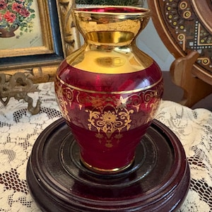 May include: A small, bulbous glass vase featuring a deep ruby red body with a reflective gold-toned neck and rim. The exterior is decorated with intricate, repeating gold filigree patterns that wrap around the center of the glass vessel.
