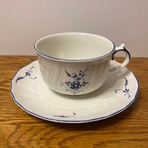 May include: A white teacup and saucer with a blue floral pattern. The cup and saucer have a scalloped edge and a blue trim.