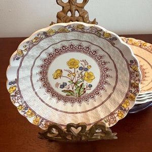 May include: A decorative plate with a scalloped edge, featuring a floral design in yellow, blue, and green on a white background. The plate is supported by a decorative metal stand. The plate's diameter is approximately 8 inches.