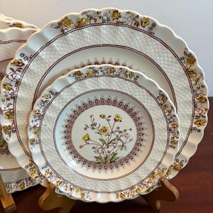 Puede incluir: Un juego de platos de cerámica blanca con un diseño floral. Los platos tienen un borde festoneado y un borde marrón con flores amarillas. Los platos están colocados sobre un soporte de madera.