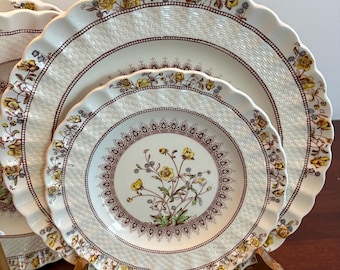 Op de afbeelding: Een set witte keramische borden met een bloemenontwerp. De borden hebben een geschulpte rand en een bruine rand met gele bloemen. De borden zijn gerangschikt op een houten standaard.