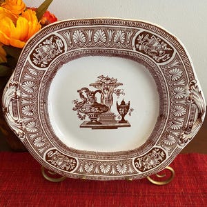 Könnte beinhalten: Ein weißer Keramikteller mit braunem Blumendesign und einem zentralen Bild einer klassischen Urne mit einer Figur, die einen Korb mit Früchten hält. Der Teller hat einen gewellten Rand und zwei Griffe.