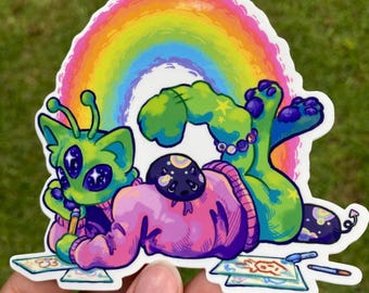 Puede incluir: Una colorida pegatina que presenta una criatura alienígena con ojos grandes, vestida con un suéter rosa y dibujando. Un arcoíris se arquea sobre la criatura. La pegatina tiene un borde blanco y la sostiene una mano.