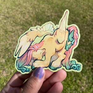 May include: A sticker featuring a yellow cartoon pony with pink and teal hair, wings, and butterfly accents. The pony is in a kneeling position with its head down. The sticker has a green border and is held by a hand.