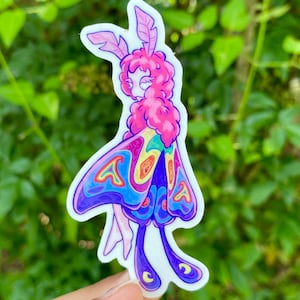 May include: A colorful sticker featuring a whimsical moth-like creature with a pink head and wings. The wings are a vibrant mix of blue, purple, yellow, and green. The sticker is held against a backdrop of green foliage.