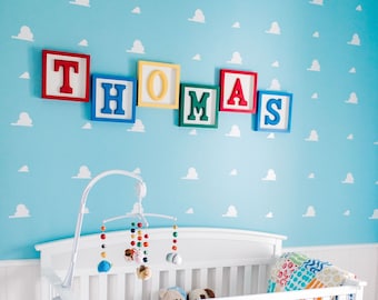 May include: A white crib with a white and grey polka dot crib skirt. The crib is decorated with a colorful mobile and stuffed toys. The wall behind the crib is painted light blue and has white cloud cutouts. The name "THOMAS" is spelled out in colorful wooden blocks above the crib.