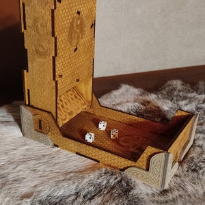Peut inclure: Une tour à dés en bois avec un motif de dragon et un motif en nid d'abeille. La tour a une fente pour que les dés roulent dans un plateau. Il y a trois dés blancs dans le plateau.