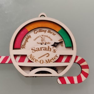 May include: A wooden Christmas bauble shaped like a candy cane with a circular gauge in the centre. The gauge is labelled "Sarah's Nice-O-Meter" and has a red, orange and green scale with the words "Naughty" and "Nice".