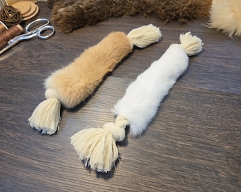 Puede incluir: Dos juguetes cilíndricos para mascotas con piel sintética suave en color canela y blanco. Cada juguete tiene una cuerda gruesa de algodón trenzado color crema en el centro, que se extiende en ambos extremos en borlas anudadas.