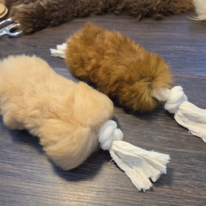 May include: Two plush dog toys. One is light tan, the other is brown. Each has a fluffy body and a white rope tassel. The toys are on a dark wood surface. Other craft supplies are in the background.