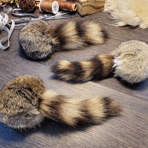 May include: Three faux fur raccoon tails with brown and tan stripes. The tails are arranged on a dark wood surface. The tails are a craft supply for costumes or other projects.