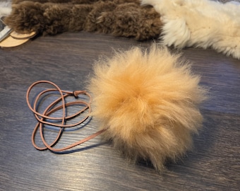 May include: A fluffy, light tan, spherical pom-pom made of soft, long-fiber material. A thin, light brown leather cord is attached to the pom-pom, forming a loose, tangled loop extending to the left side of the round, textured object.