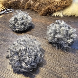 May include: Three gray, curly wool balls are arranged on a wooden surface. The wool has a natural, textured appearance. The background includes other wool pieces and crafting tools, suggesting a handmade craft or textile project.