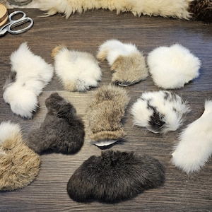 May include: A collection of fluffy, textured fur pieces in various colors, including white, gray, brown, and black. The pieces are arranged on a dark wooden surface, showcasing the natural variations in texture and color.