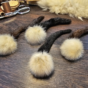 May include: Four fluffy white pom-poms with brown, black, and gray fur tails. The pom-poms are on a wooden surface.