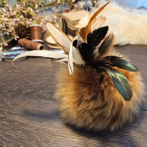May include: A fluffy brown pom-pom with a variety of feathers attached. The feathers are white, brown, black, and green.