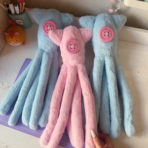 Peut inclure: Trois peluches pieuvres, deux bleues et une rose. Chacune a un bouton sur la tête. La pieuvre rose est au centre, flanquée de deux bleues. Les jouets ont de longs tentacules doux. Une main avec du vernis à ongles blanc est visible.