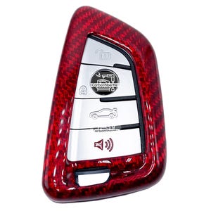 May include: A red carbon fibre car key fob cover with silver buttons. The key fob has a textured, glossy finish and features icons for lock, unlock, trunk, and alarm. The text "Carbon Fiber Me" is visible on the key fob.