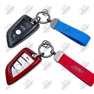 Puede incluir: Dos llaveros de coche. Uno es negro con un diseño de fibra de carbono, el otro es rojo con un diseño de fibra de carbono. Ambos tienen un anillo de metal y una correa. Una correa es azul con un logotipo, la otra es roja con un logotipo.