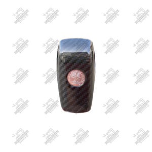 May include: A black carbon fiber car shift knob with a chrome top and a copper-colored emblem. The shift knob has a textured, woven pattern and a rounded rectangular shape.