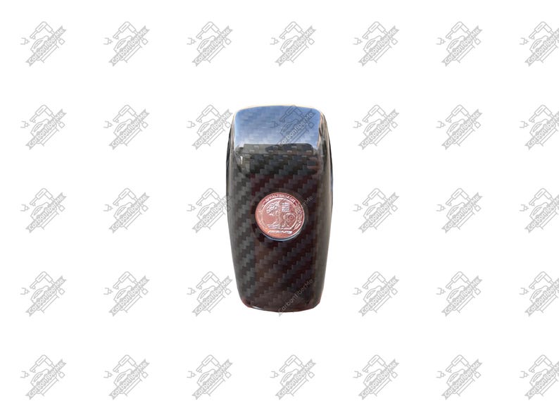 May include: A black carbon fiber car shift knob with a chrome top and a copper-colored emblem. The shift knob has a textured, woven pattern and a rounded rectangular shape.