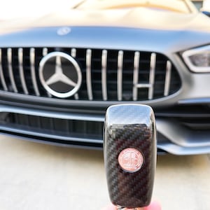 May include: A close-up of a car key with a carbon fiber design and a circular emblem. The key is held in front of a gray Mercedes-Benz car, with the front grille and emblem visible in the background.