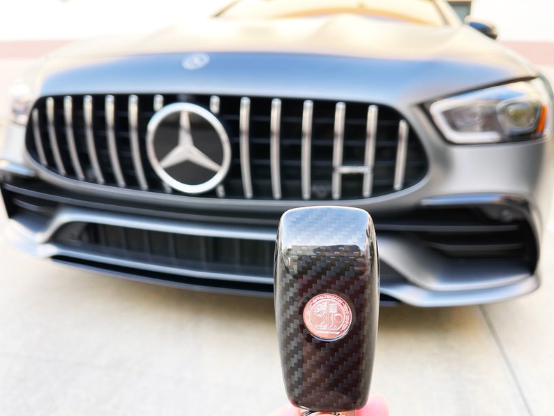 May include: A close-up of a car key with a carbon fiber design and a circular emblem. The key is held in front of a gray Mercedes-Benz car, with the front grille and emblem visible in the background.