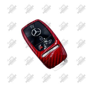 May include: Red carbon fiber car key fob cover with black buttons and the Mercedes-Benz logo. The key fob has the AMG logo and other symbols. The key fob is a car accessory.