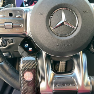 May include: Close-up of a black leather steering wheel with silver accents. The Mercedes-Benz logo is centered, with an airbag label below. Features include cruise control buttons, a carbon fiber gear shift, and an AMG emblem.
