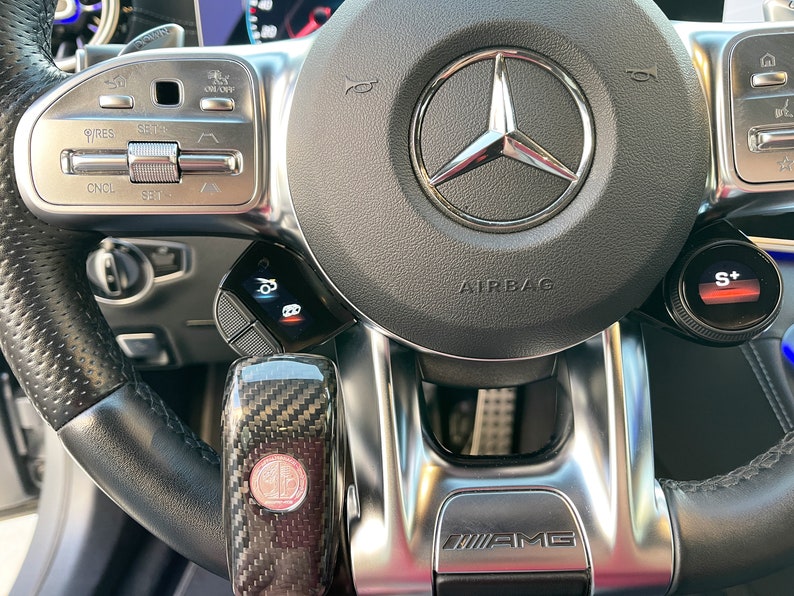 May include: Close-up of a black leather steering wheel with silver accents. The Mercedes-Benz logo is centered, with an airbag label below. Features include cruise control buttons, a carbon fiber gear shift, and an AMG emblem.