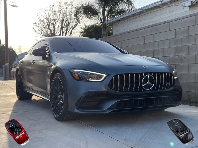 May include: A gray luxury car with a black grille and black wheels is parked on a concrete driveway. The car has the Mercedes-Benz logo on the front. Two car keys are in the foreground.