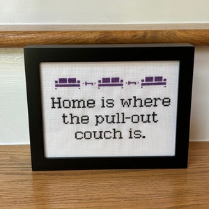 May include: A framed cross-stitch with a purple couch design and the text "Home is where the pull-out couch is."