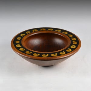May include: A round wooden bowl with a smooth, dark brown finish. The wide, flat rim features a black inlay band decorated with a repeating pattern of small, yellow, spiral-shaped motifs arranged evenly around the entire circumference.