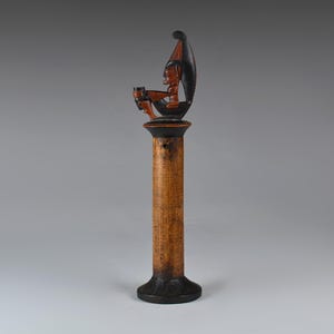 Puede incluir: Una escultura de madera tallada que representa una figura sentada en una estructura en forma de barco sobre una columna. La figura tiene piel de color marrón rojizo y un tocado. La columna es de color marrón claro con una base y una parte superior oscuras. Es probable que sea un objeto decorativo.