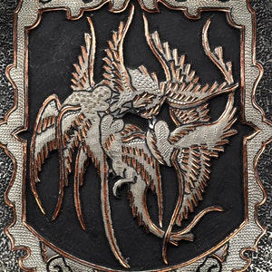 May include: A silver and copper metalwork design of two birds in flight, with intricate details and a black background.