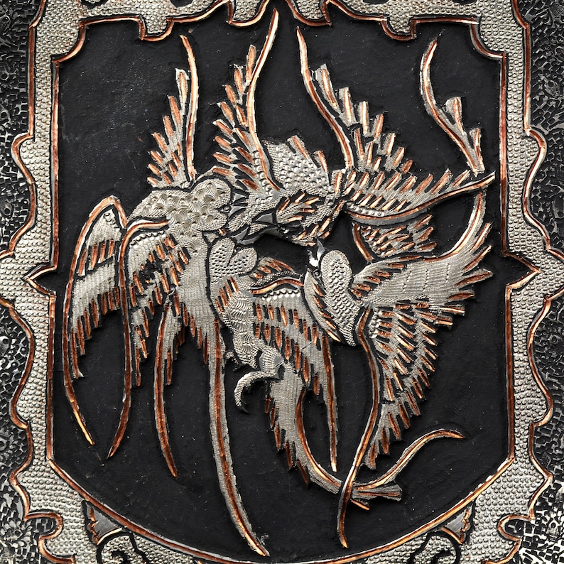 May include: A silver and copper metalwork design of two birds in flight, with intricate details and a black background.