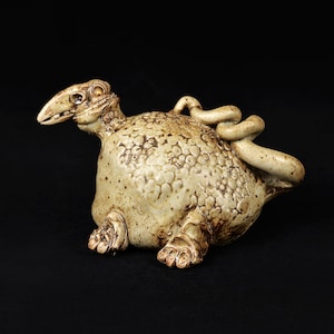 Könnte beinhalten: Beige Keramikskulptur einer fabelhaften Kreatur mit reptilienartigem Kopf, strukturiertem Körper und eingerolltem Schwanz. Die Skulptur hat braune Akzente und steht auf schwarzem Grund.