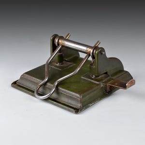 May include: A vintage green metal hole punch with a lever and a metal base. The punch has a worn, aged look.