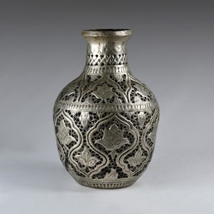 May include: A silver-toned metal vase featuring an intricate openwork lattice design. The surface is decorated with embossed floral motifs arranged within arched panels, complemented by a textured neck and a narrow, circular opening at the top.
