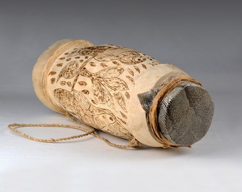 May include: A cylindrical drum with a natural, light-coloured finish. The drum features etched bird and leaf designs. The ends are covered with a mesh material and secured with a woven band. A rope is attached for carrying.